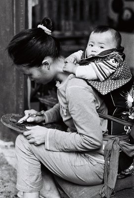 A young mother is carving souvenirs for tourists, her child tied on her back in the old town of Lijiang, Yunnan.