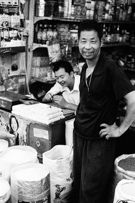 Standing amidst his goods, a trader in rice, beans, oil and other canned goods smiles at the camera. Their stall was stuffed to the rafters with bottles and cans, sacks of rice and drums full of oil. The small counter features a poster with a modern, beautiful woman, electronic scales and a small assortment of noodles.

Set to one side of the market, these stall were fun to see and made for interesting discoveries of goods and people alike.