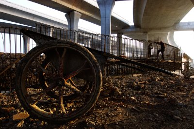 An old rusty wheel barrow sits in a field of debris. The morning sun lights it up, as in the distance workers are braiding steel for another piece of high way. 

Overhead several highways are parting ways . I like the juxtaposition of the massive highways and the simple, human powered wheel barrow. Sights like these are still very common in China and it always makes you wonder what could be achieved with better tools.