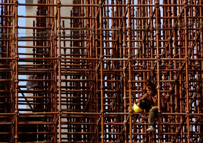 A young worker is taking five high up in the steel scaffold on a high way construction. The early morning sun casts a nice light on him. The steel rods and his clothes are the same color, the only break is the yellow helmet. 

He looks lost, deep in thought, maybe wondering how much more construction there will be in his lifetime.