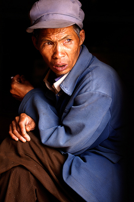 An older peasant is sitting in on the door step of a restaurant, smoking cigarette stubs given to him by the patrons. 
This image presented itself to me when I was having lunch in the old town of Shaxi in the mountains of Yunnan. The man slowly inched closer the our table the other patrons, gathering cigarette stubs and also hoping for something to eat.

The village is in a remote part of Yunnan, with few tourists and the farmers come from the surrounding villages to market every week. They are easy targets for the locals, since they hardly know the value of money and live a very secluded live.

The image was shot as he was sitting in the door, bathed in the light of the day against a dark interior.